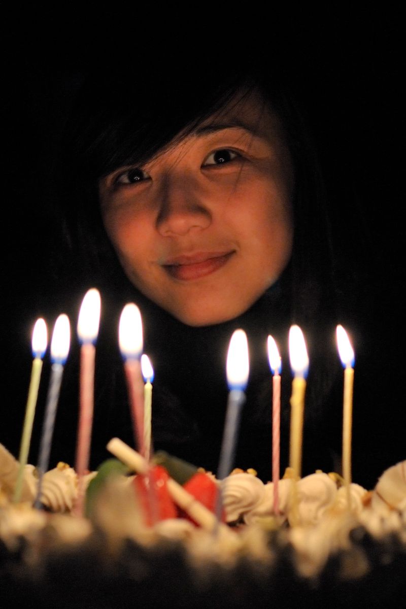 A woman smiling behind a birthday cake with lit candles in a dimly lit room, creating a warm and festive atmosphere.