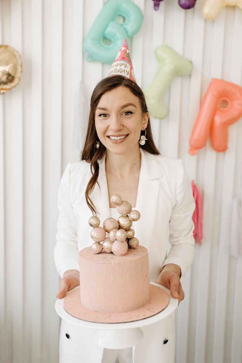 Smiling woman in party hat holds a pink birthday cake at a celebration indoors.